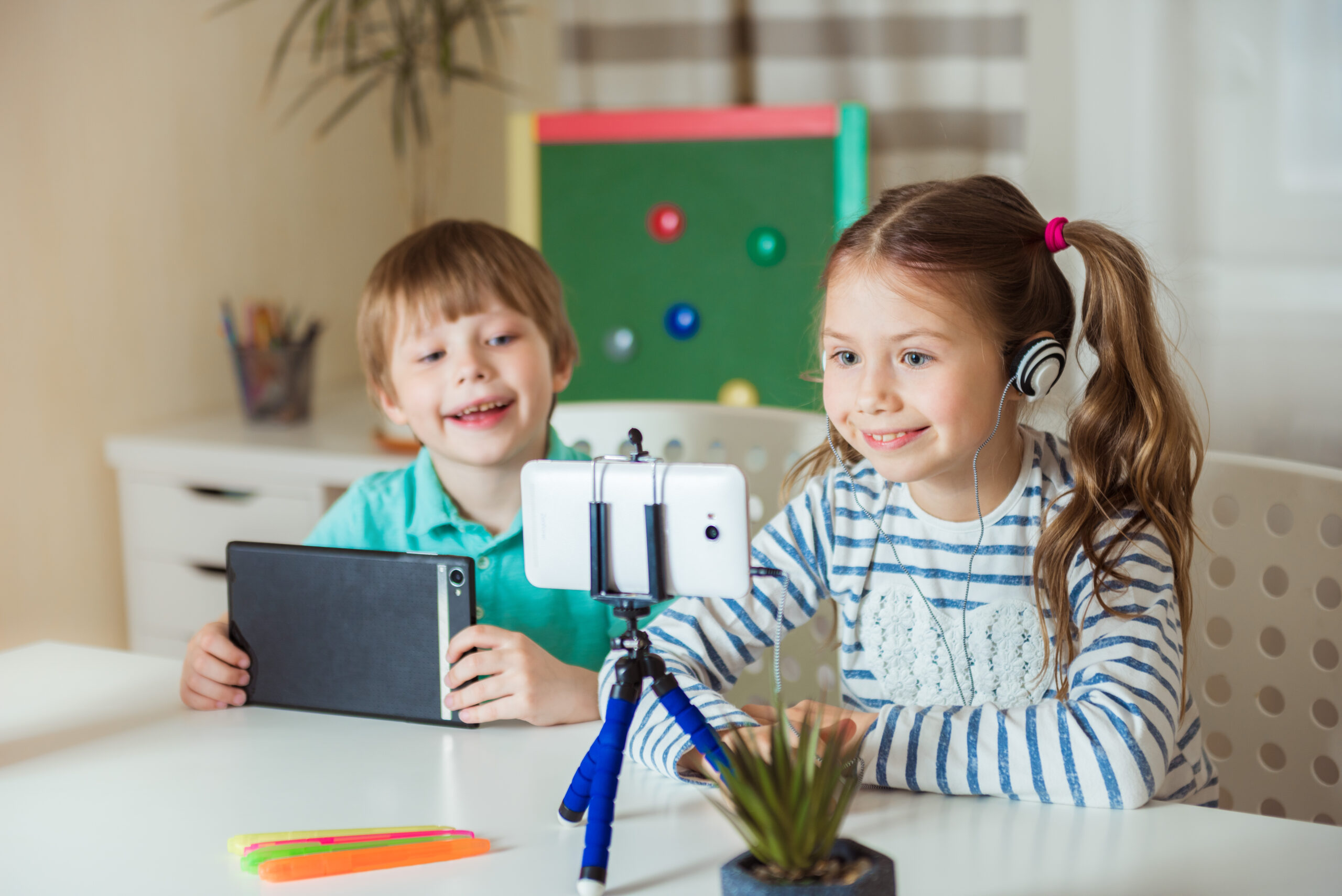 Niño y niña pequeños participando en una videollamada educativa, con auriculares, tableta y un teléfono móvil sobre trípode en una mesa.