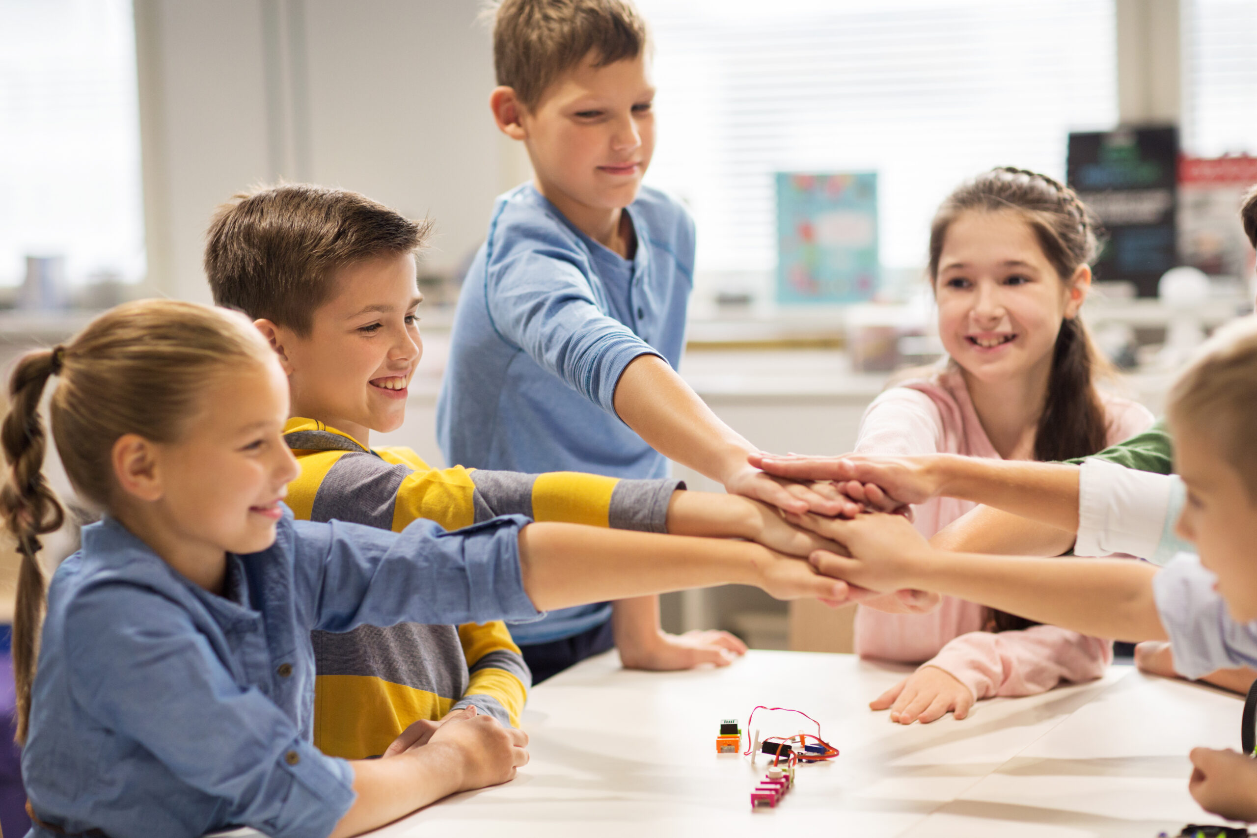Grupo de niños en un aula juntando las manos en el centro de la mesa como símbolo de trabajo en equipo.