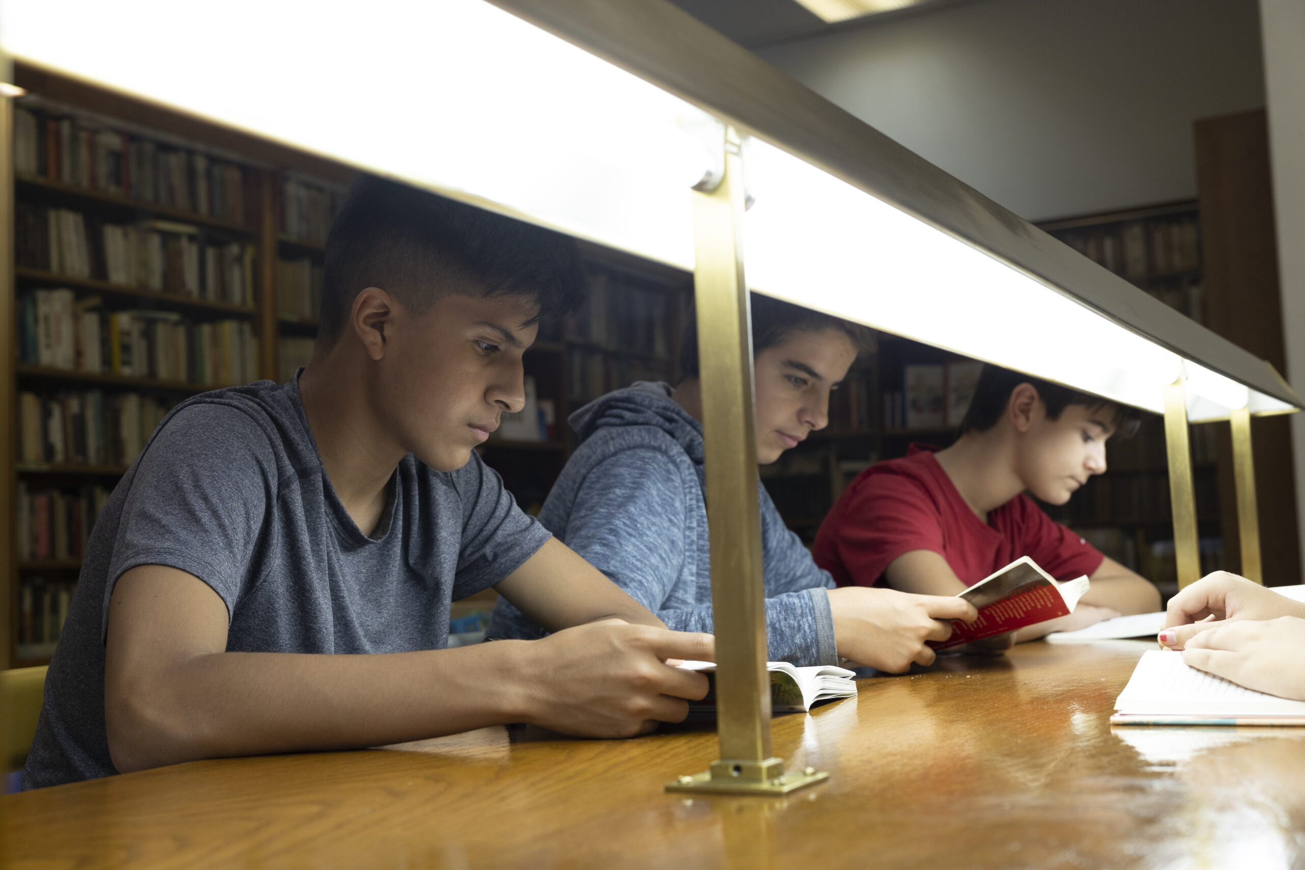 Tres adolescentes leyendo y estudiando en una biblioteca, sentados bajo una lámpara de mesa.