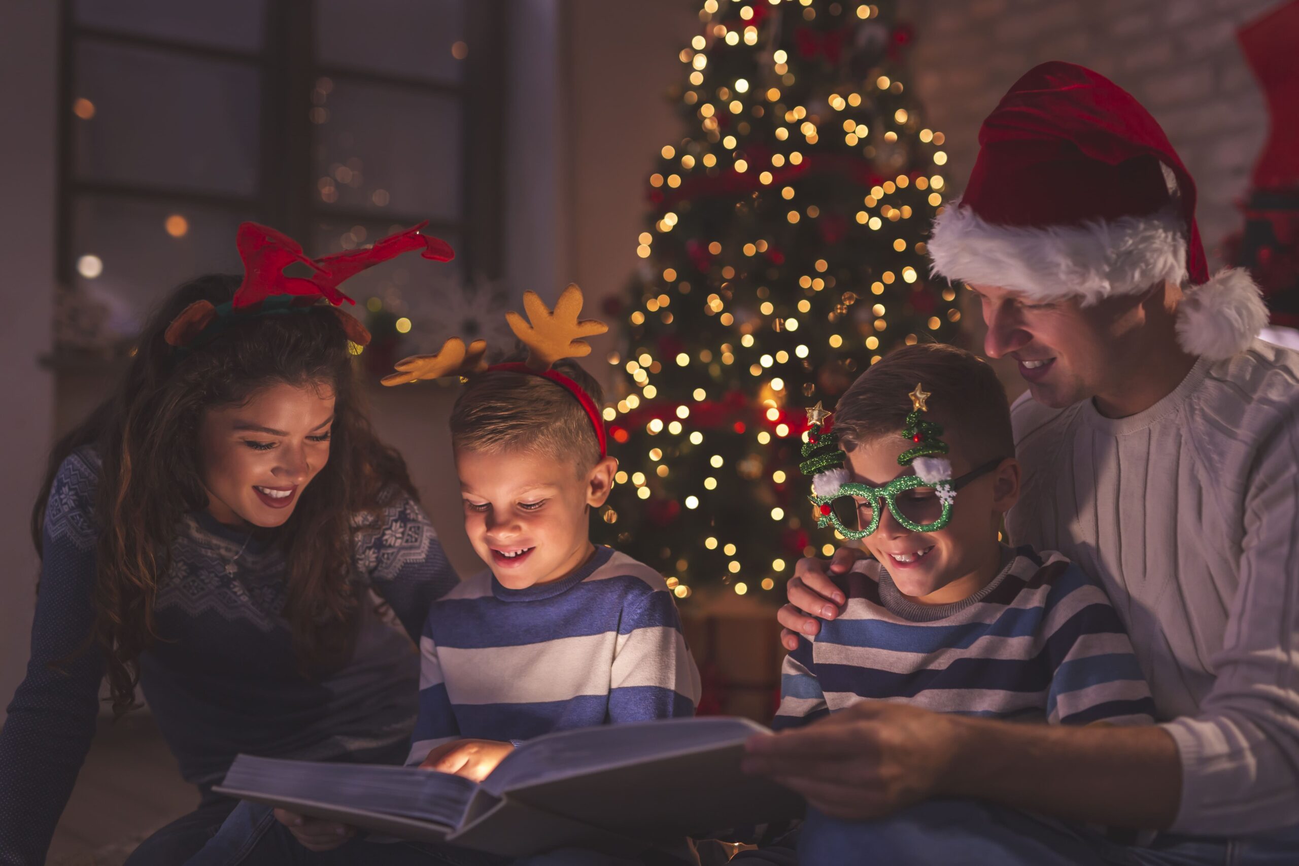 Familia leyendo un cuento de Navidad con niños junto al árbol, compartiendo una actividad educativa navideña en casa.
